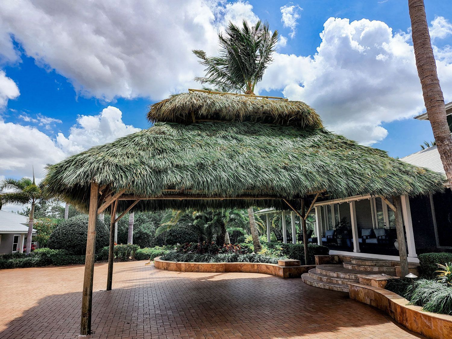 Thatched-roof carport with brick pavers, wooden posts, and manicured landscaping under a cloudy sky.