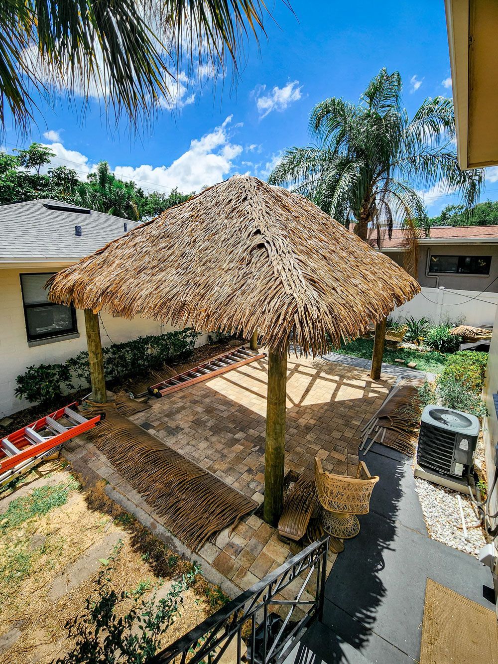 Tiki-style roof over a brick patio with wooden posts. Lush greenery surrounds the structure under a bright sky.