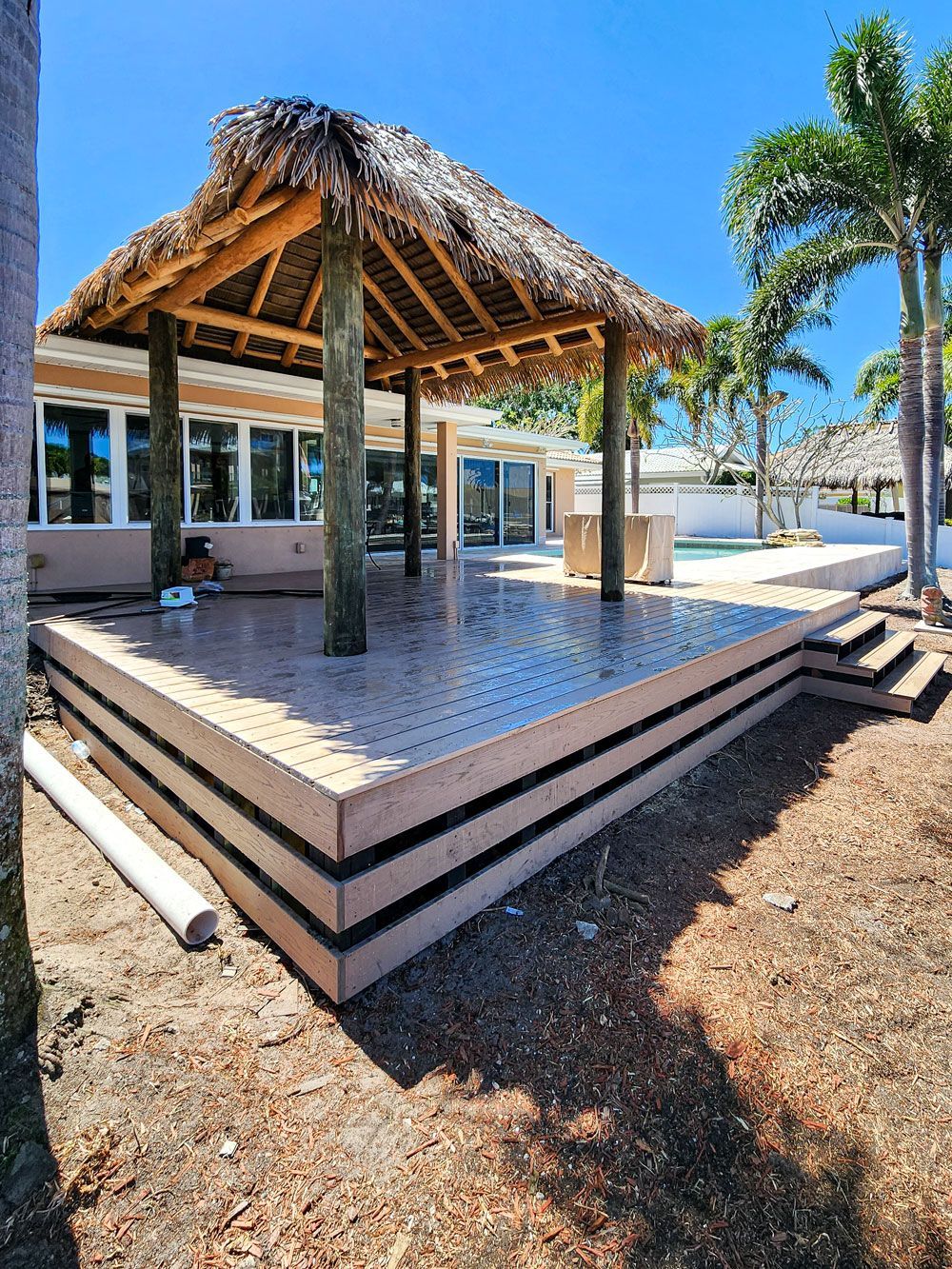 A thatched-roof gazebo on a raised platform, next to a building and palm trees, under a bright blue sky.