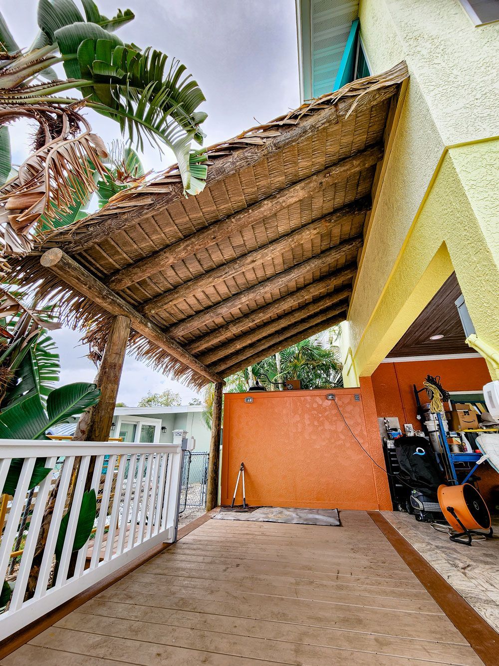Thatched roof extending from a yellow stucco building, overlooking a patio with a white fence.