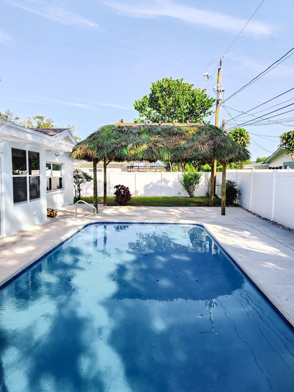 Backyard with a pool covered in blue, a tiki hut, and white fence under a sunny sky.