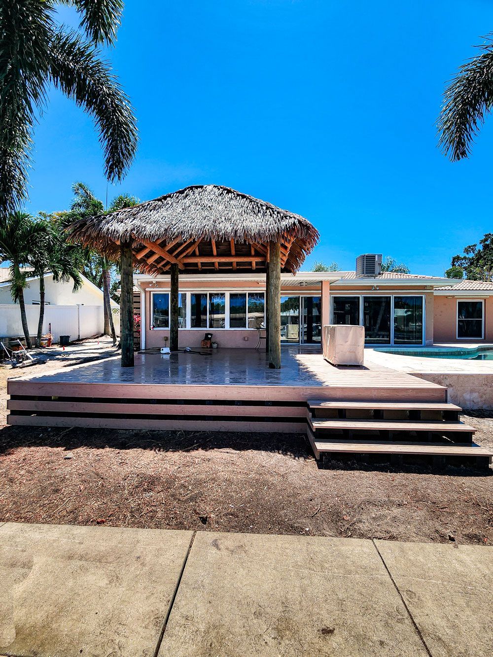 Gazebo with thatched roof on a raised patio, near a house with a pool, under a blue sky.