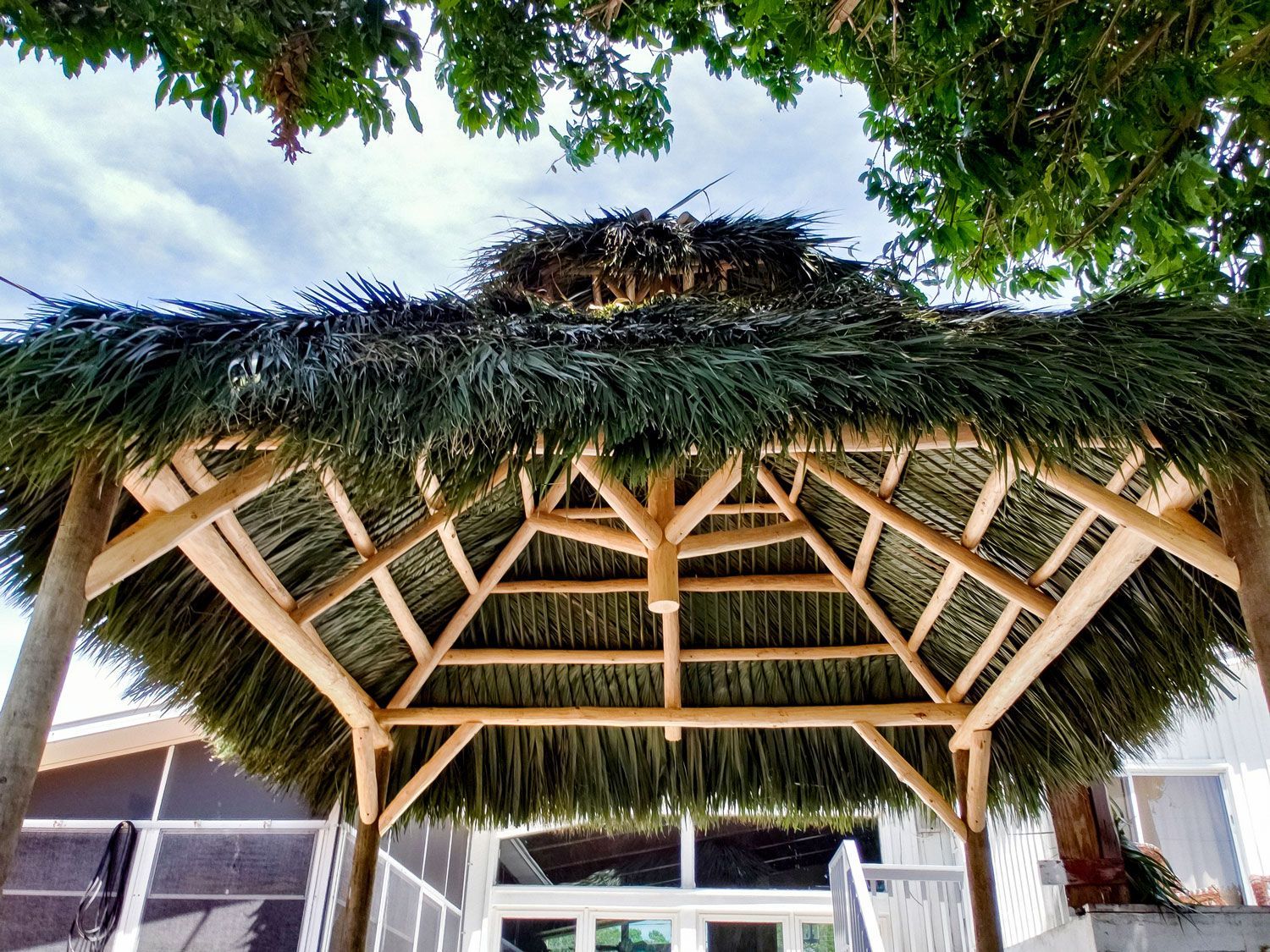 Tiki hut with a thatched roof, wooden beams, and greenery against a blue sky.