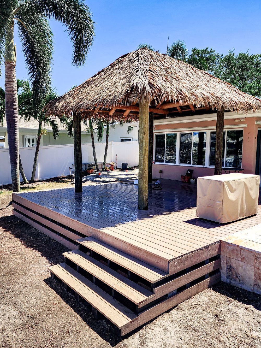 Thatched-roof gazebo on a wooden deck with steps, in a yard. Sunny day, with palm trees and a building in the background.