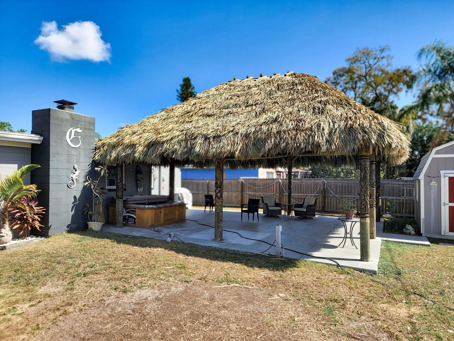 Thatched roof patio with seating, grill, and shed on a sunny day.