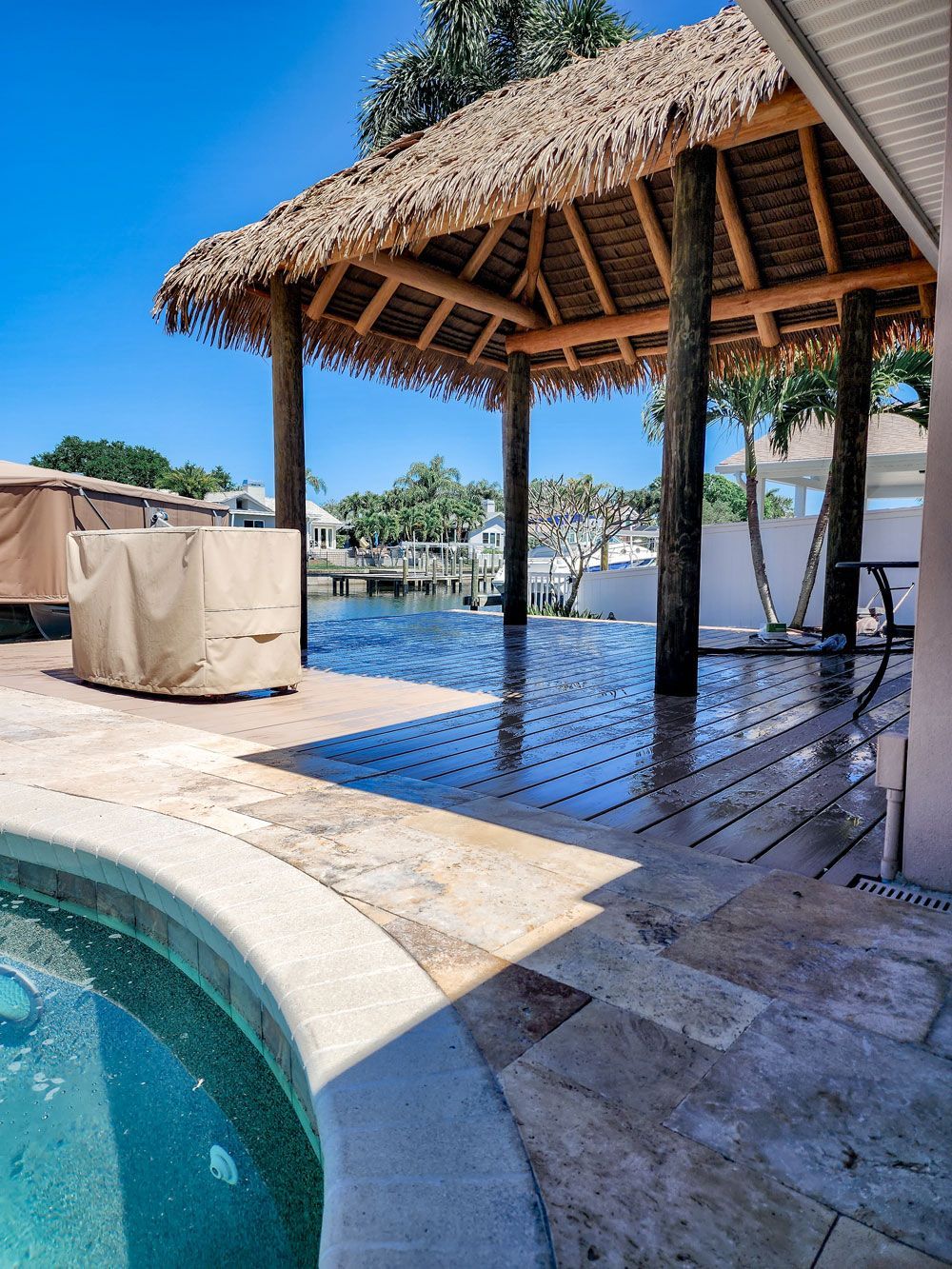 Poolside tiki hut overlooking canal. Pool, covered patio, blue sky, water, and palm trees.