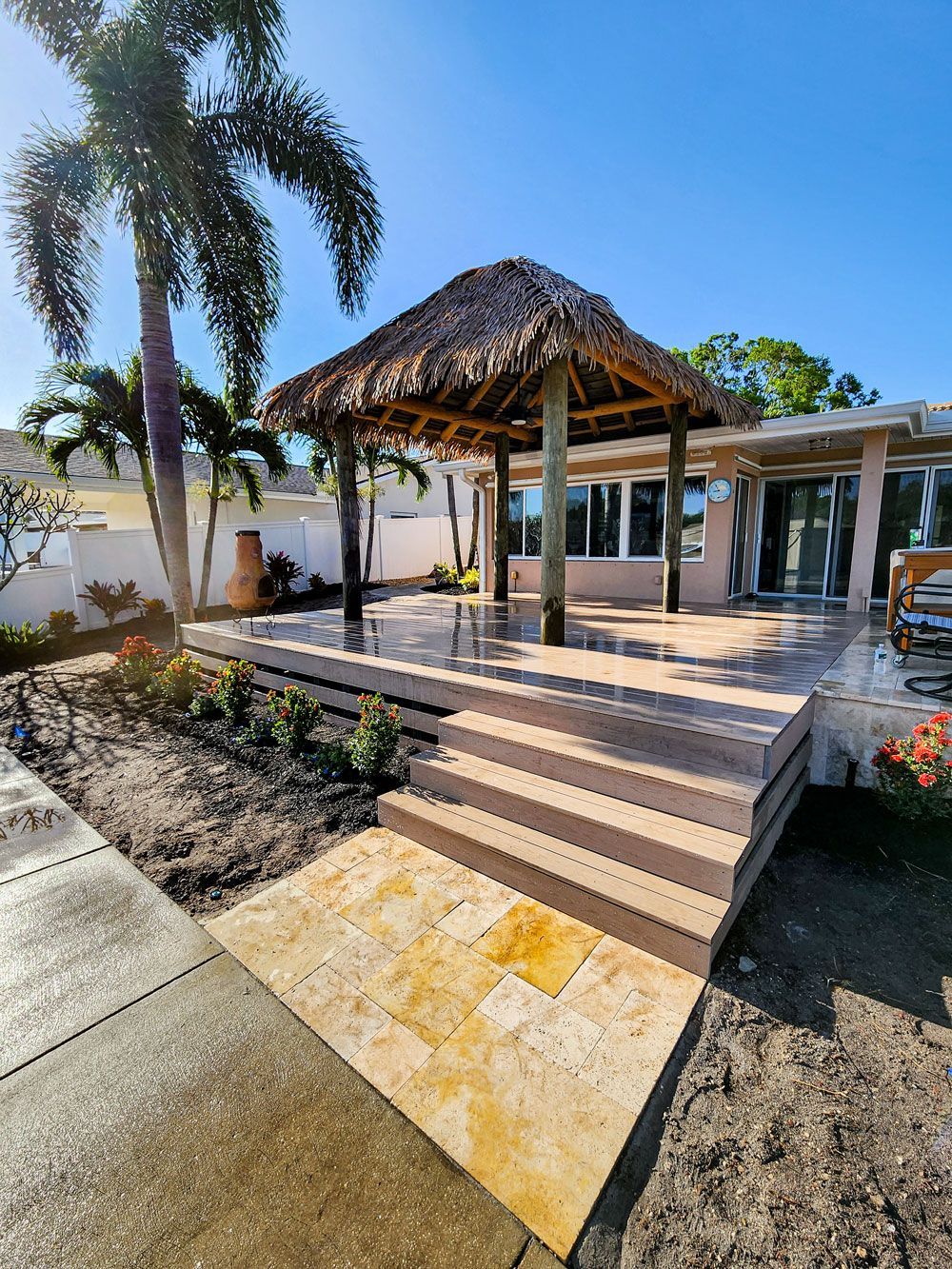 Tropical-style gazebo with a thatched roof, on a raised deck with steps, near a house and palm trees. Sunny day.