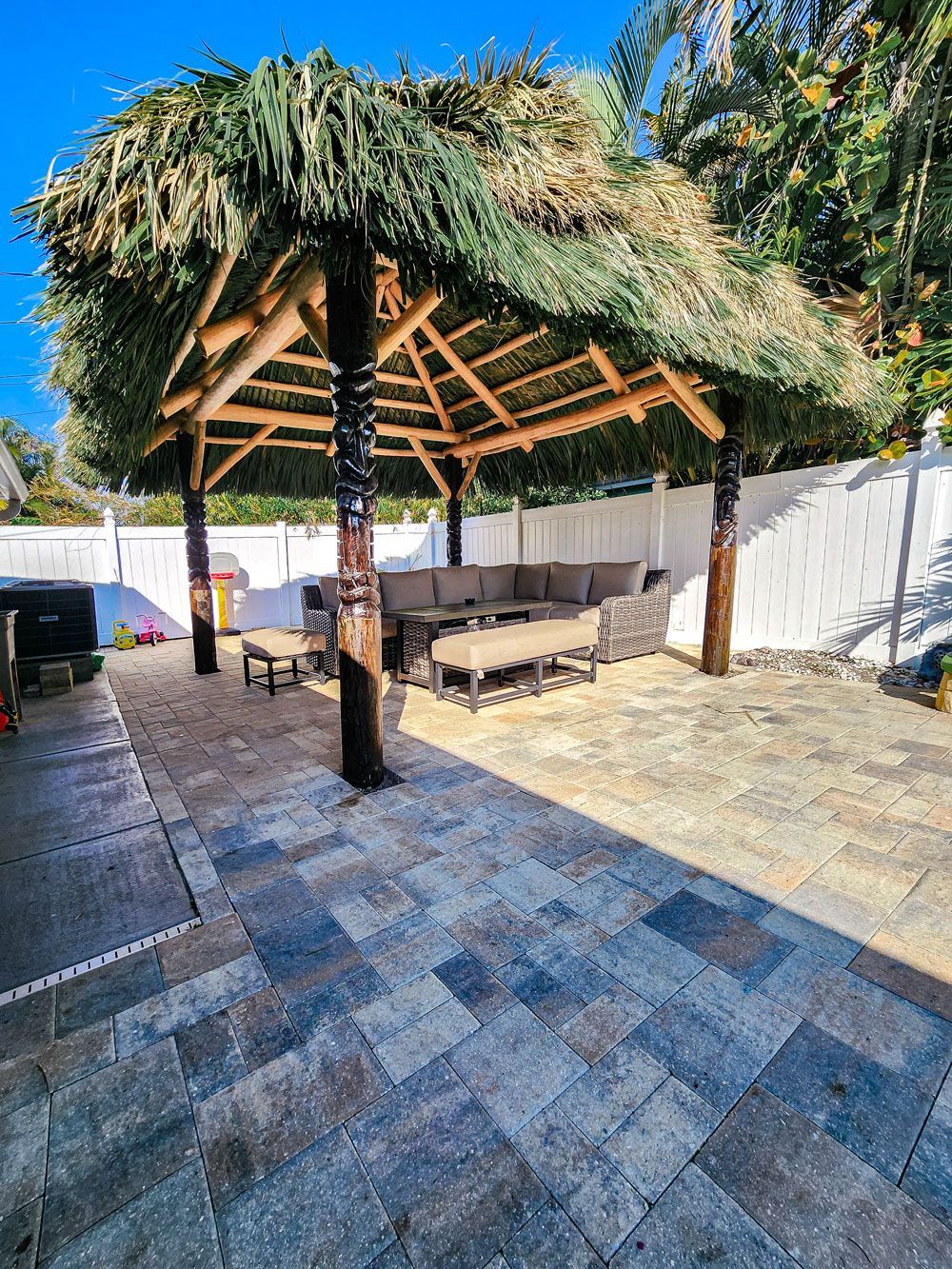 Outdoor thatched-roof gazebo with seating on a brick patio in a backyard with a white fence.