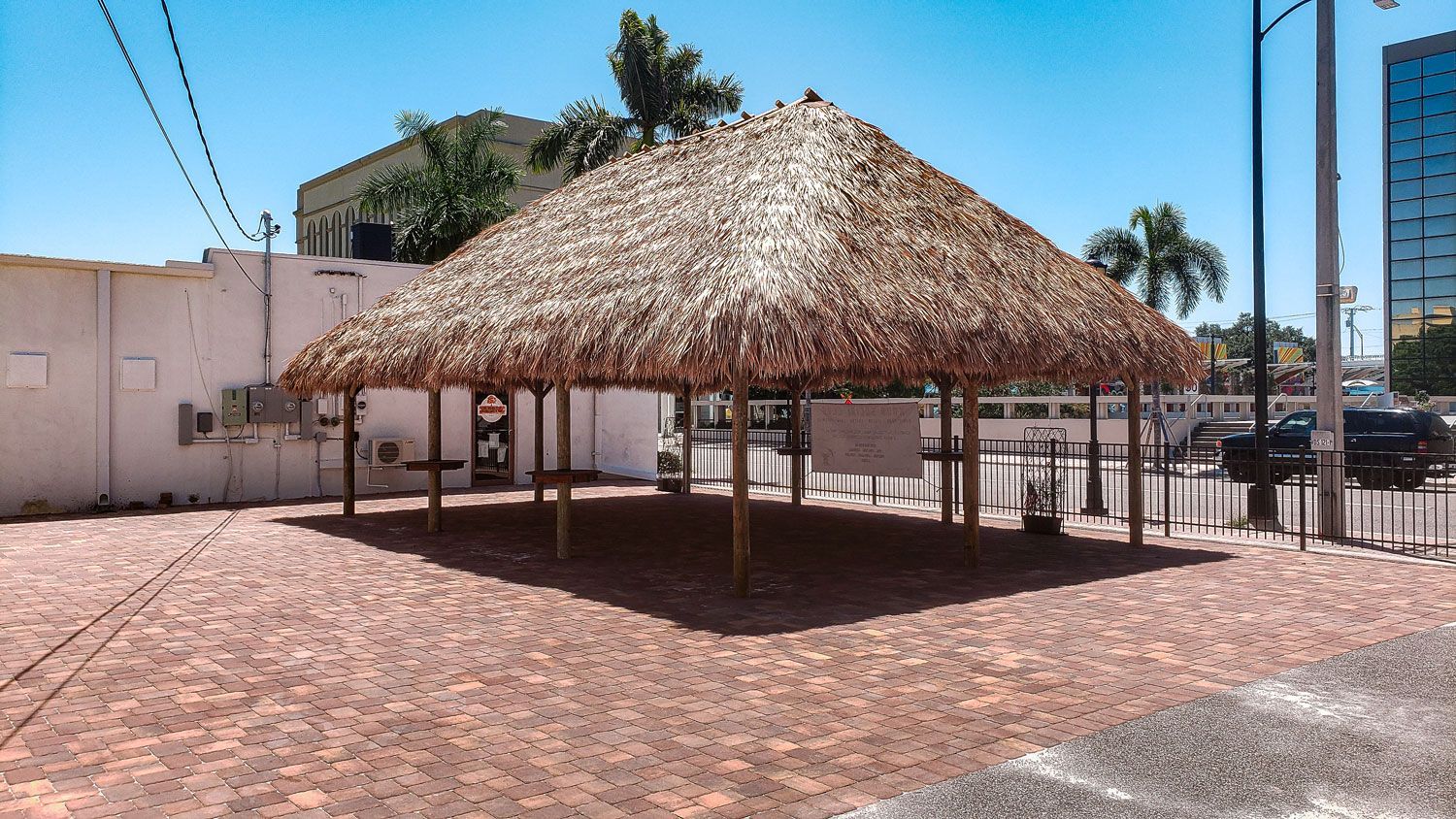 Thatched-roof outdoor shelter on brick pavers, with surrounding buildings and blue sky.