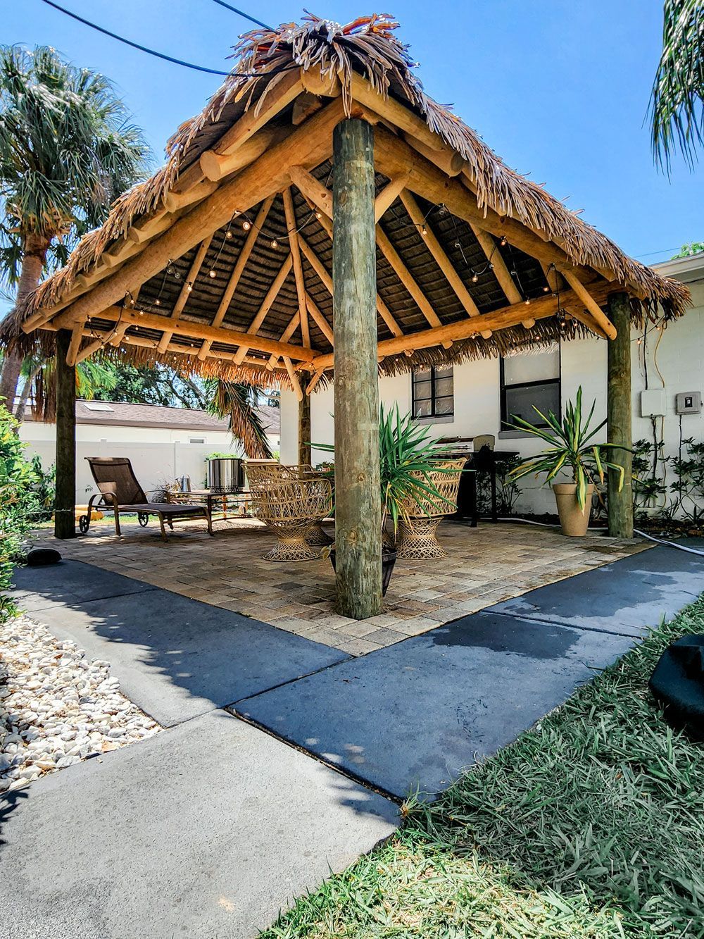 Tiki hut with thatched roof and wooden supports over a gravel patio. Concrete pathway leads to it.