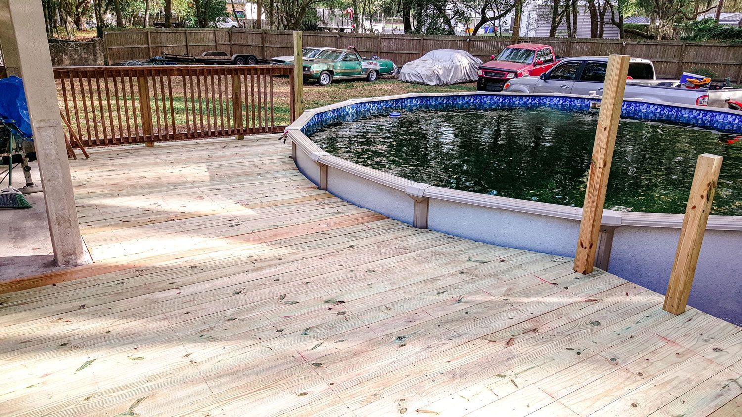 Wooden deck surrounds a round pool with green water. Cars parked in the background.
