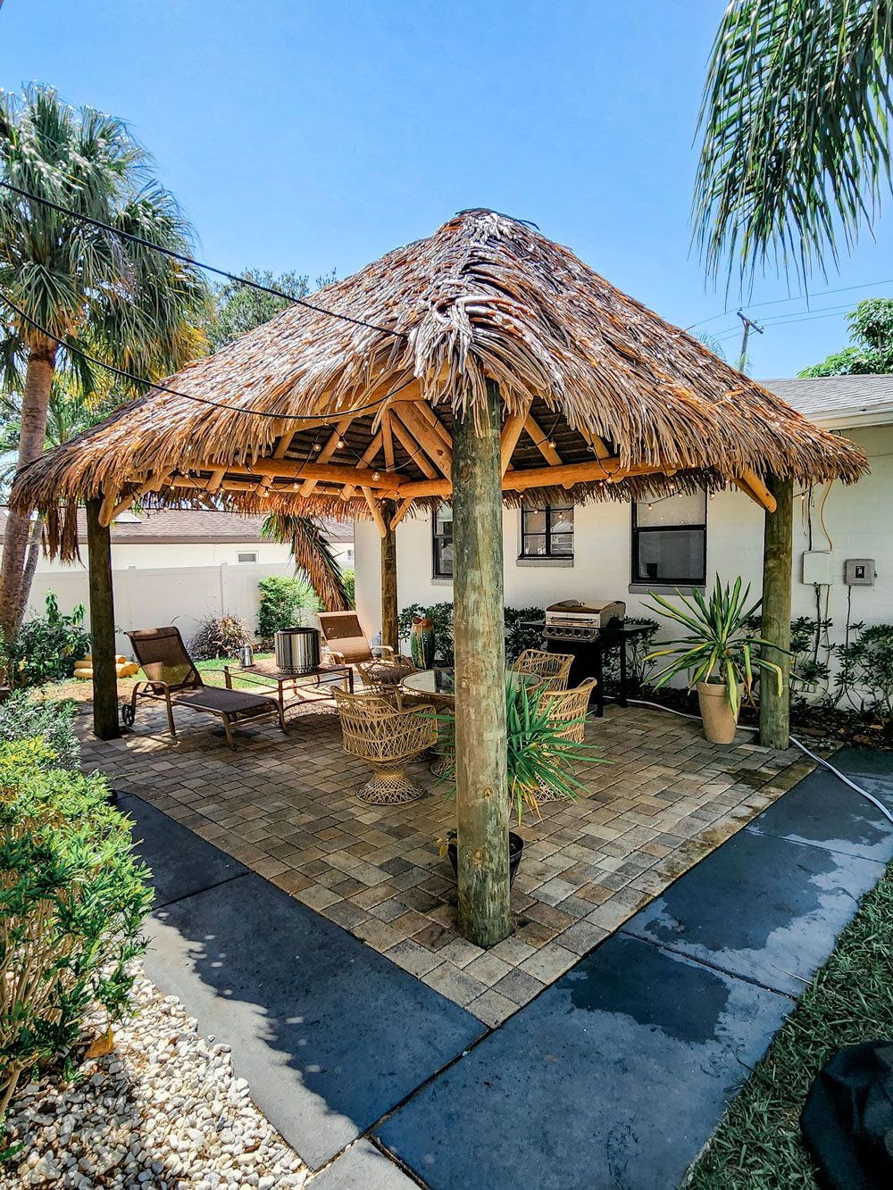 Tiki hut over brick patio with seating and grill near a house.