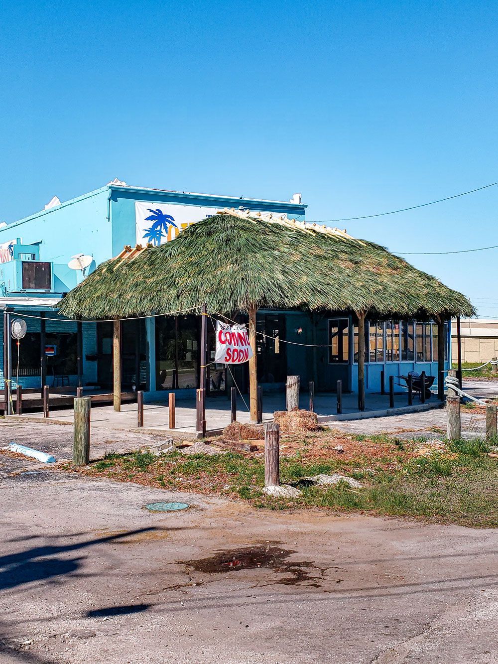 Tiki-style restaurant with a thatched roof, blue building, and palm tree sign against a blue sky.