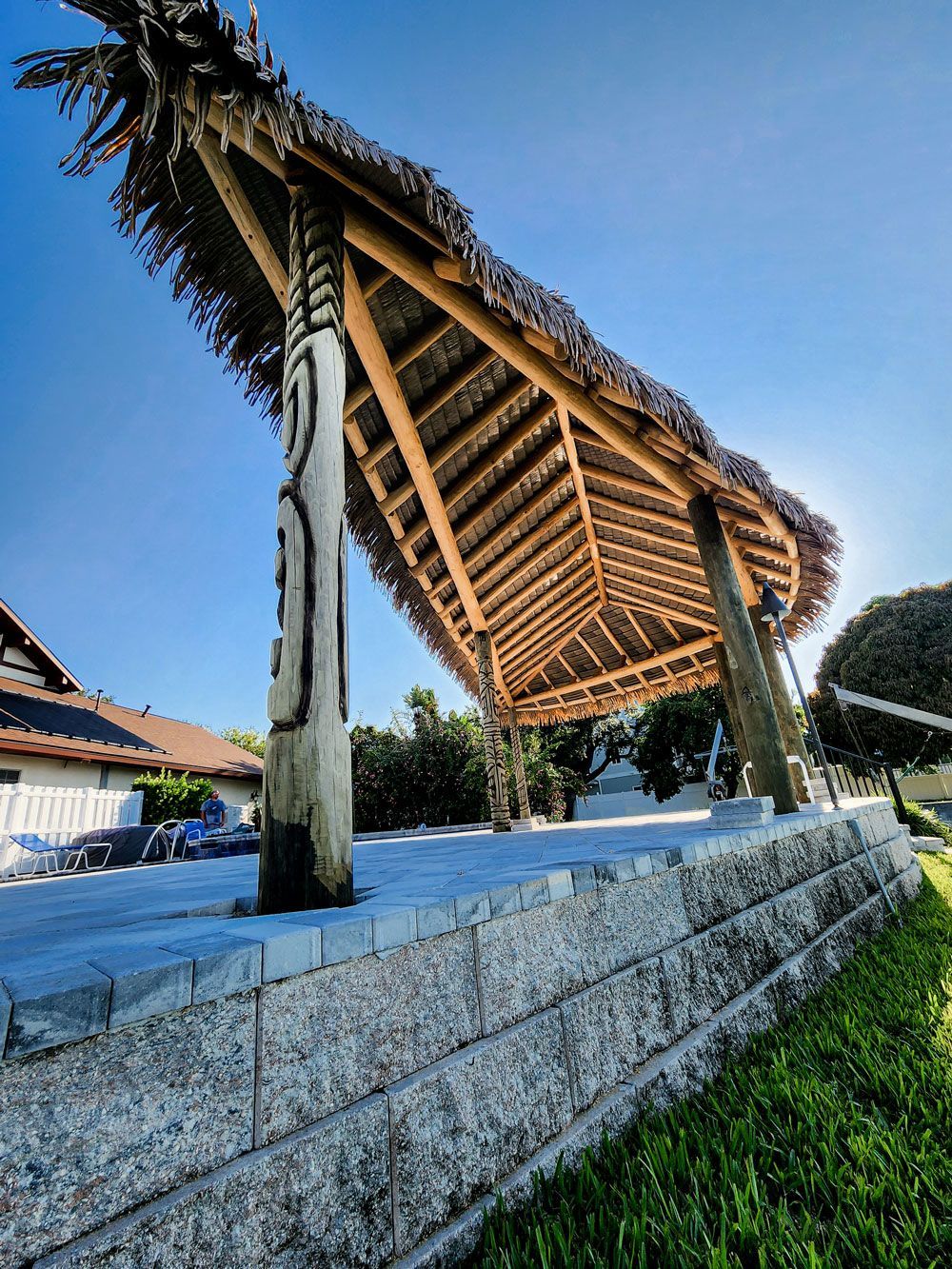 Tiki-style shelter with thatched roof, wooden posts, on a stone platform, blue sky background.