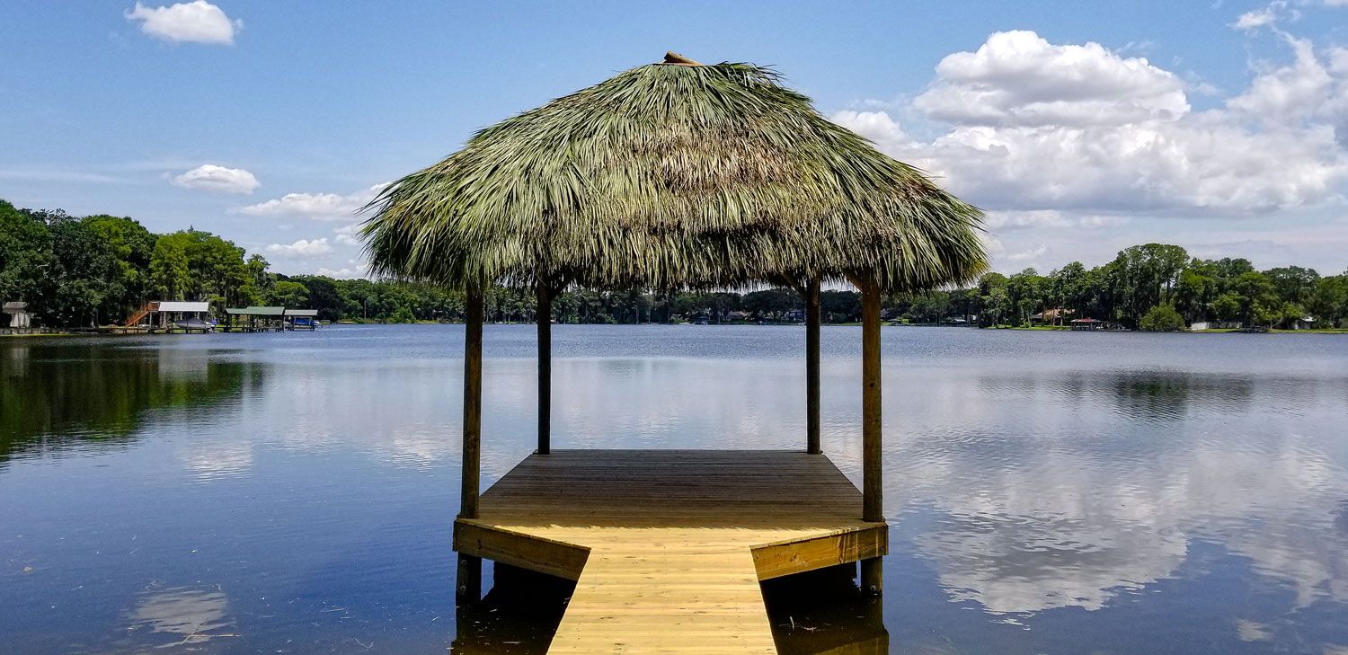 A thatched-roof gazebo on a wooden dock over calm water reflects the blue sky and clouds.