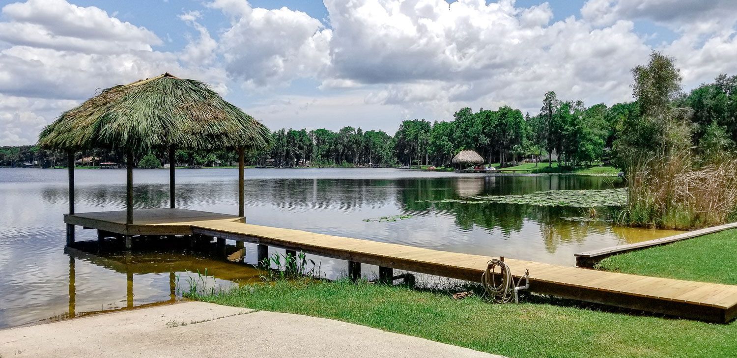 A thatched-roof dock extends over calm water on a sunny day. Trees and clouds fill the horizon.