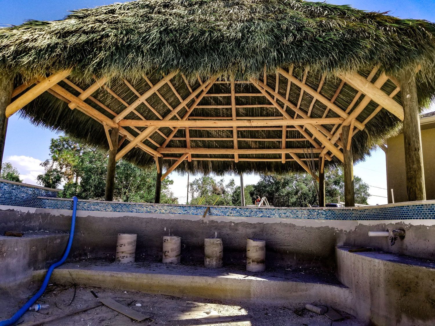 Palapa with thatched roof over a pool under construction; blue hose and cement supports visible.