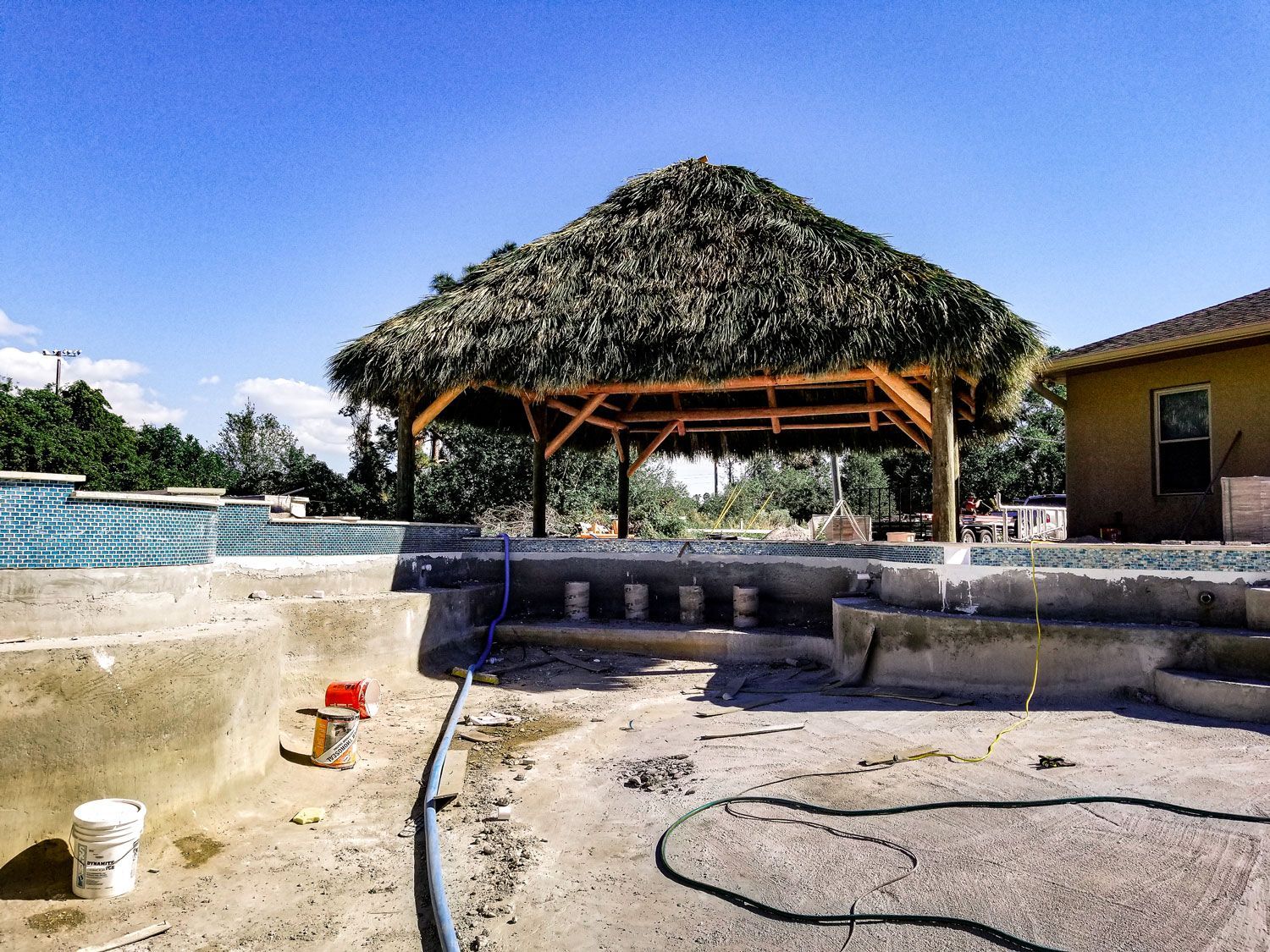 Pool under construction with thatched roof shelter; blue sky.