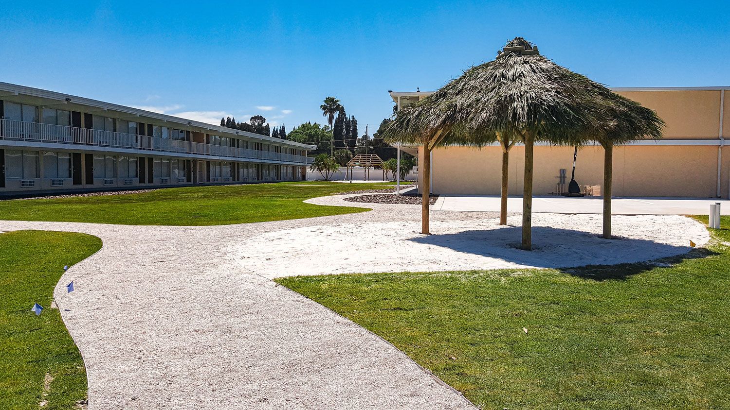 Exterior view of a building with a grassy lawn, white gravel path, and a thatched-roof shelter under a blue sky.