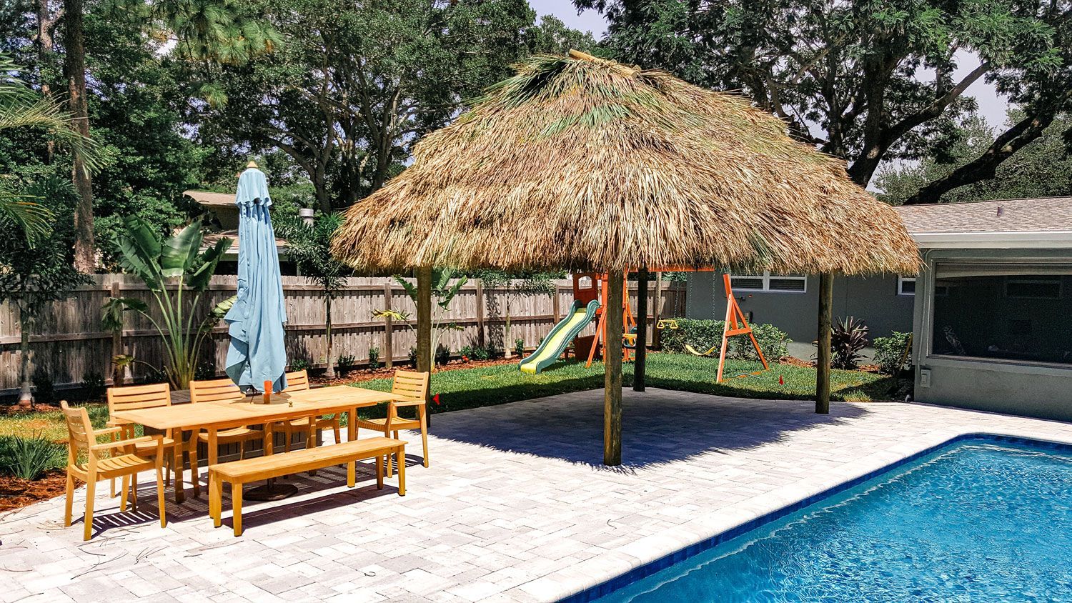 Tiki hut with thatched roof over a patio with dining set and a pool.