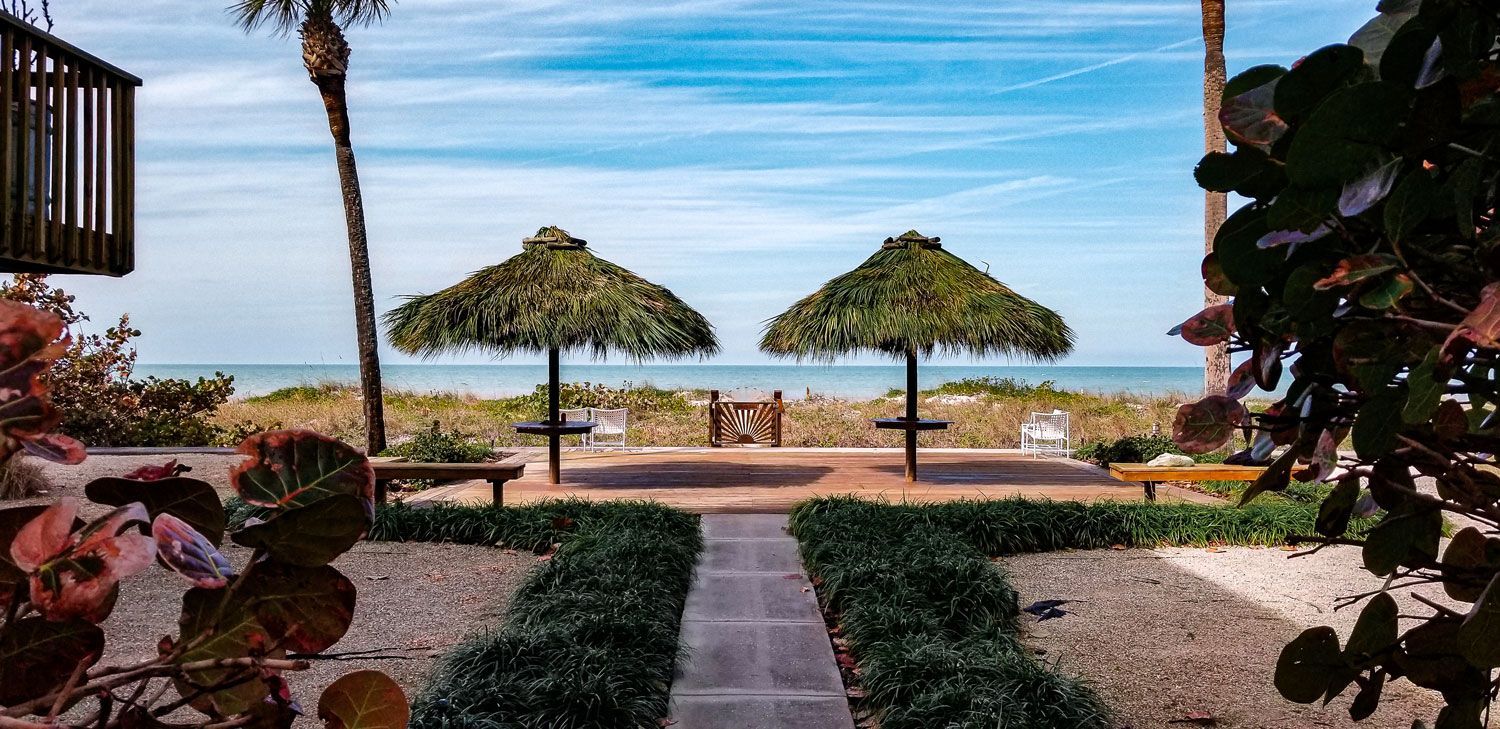 Two thatched umbrellas on a beach pathway, leading to the ocean. Blue sky, greenery, and benches.