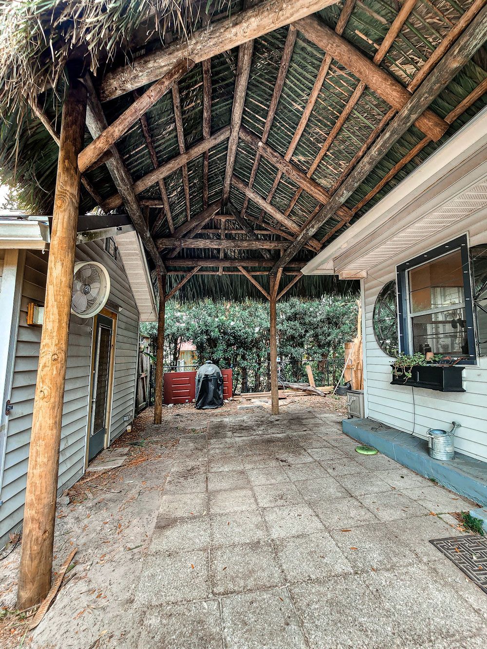 Covered outdoor patio with a wooden frame and thatched roof. Concrete flooring. Adjacent to a house and shed.
