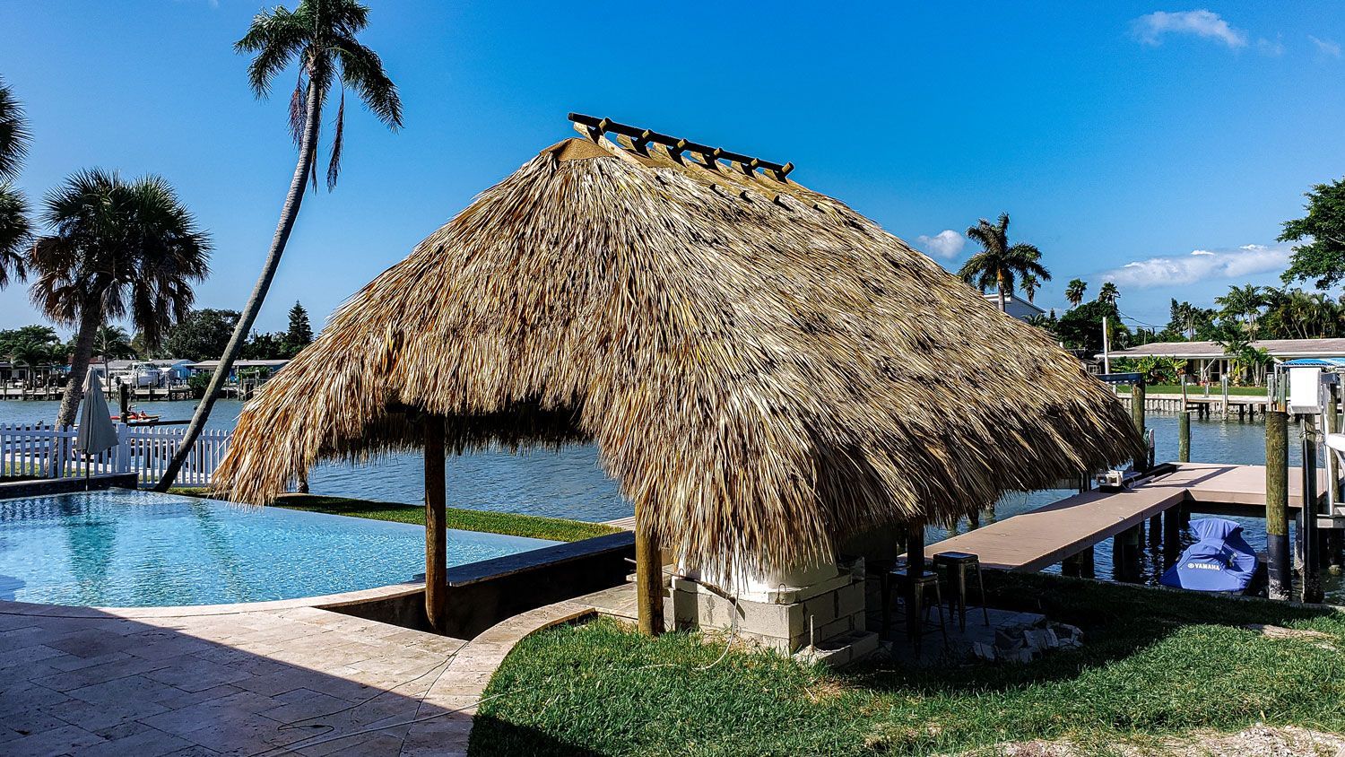 Thatched-roof tiki hut beside a blue pool and a wooden dock, with palm trees and a sunny sky.