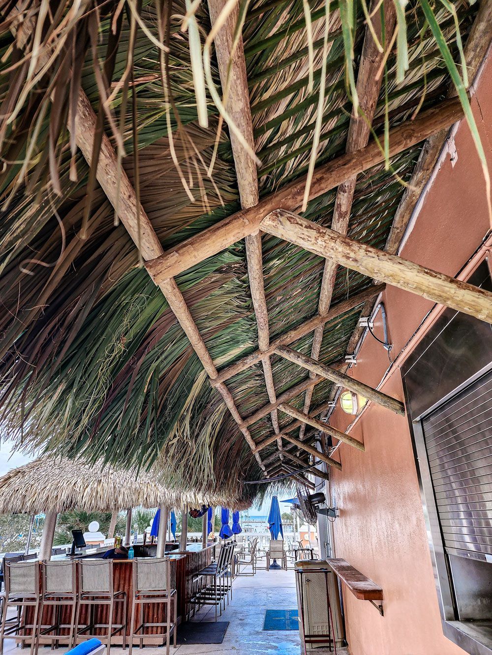 Tiki hut roof over a bar area with tables, blue umbrellas, and a beach setting.