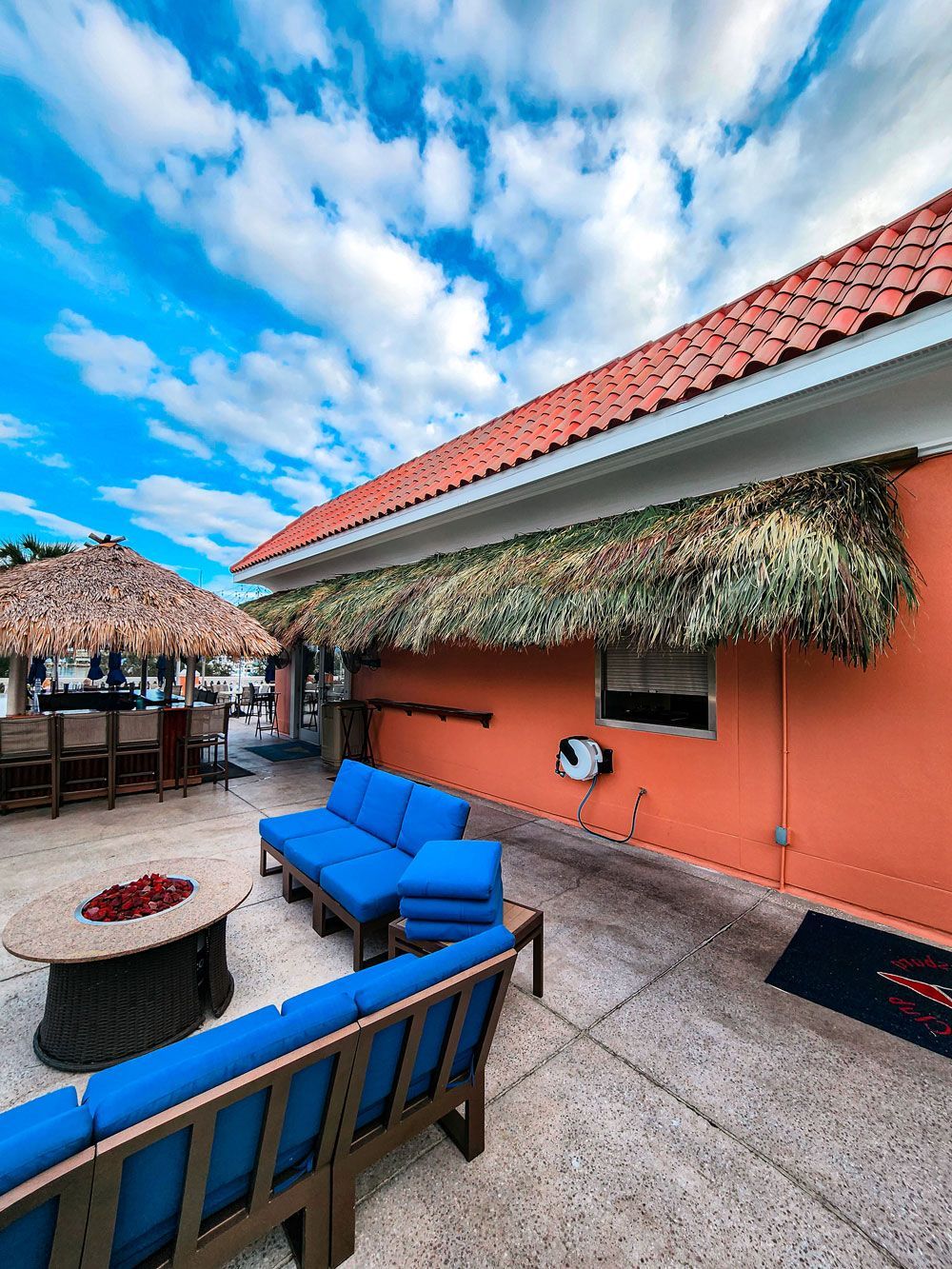 Outdoor patio with blue couches, a fire pit, and a tiki bar under a blue sky.