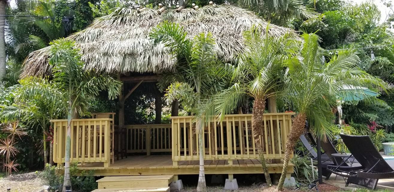 Tropical-style gazebo with thatched roof, surrounded by palm trees and greenery; poolside.