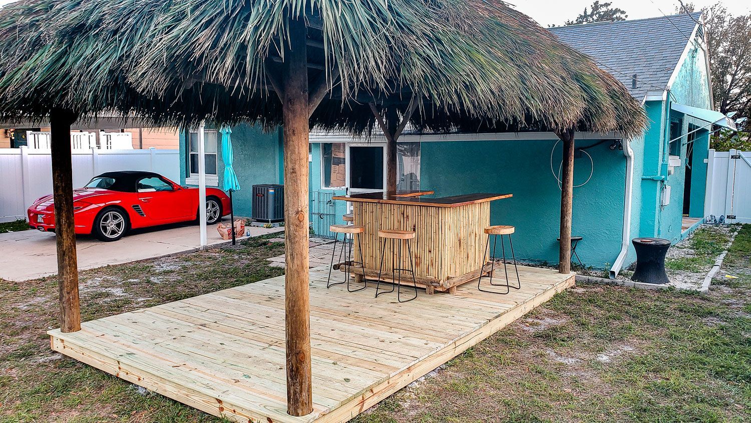 Outdoor tiki bar with thatched roof, wooden deck, and a red sports car.