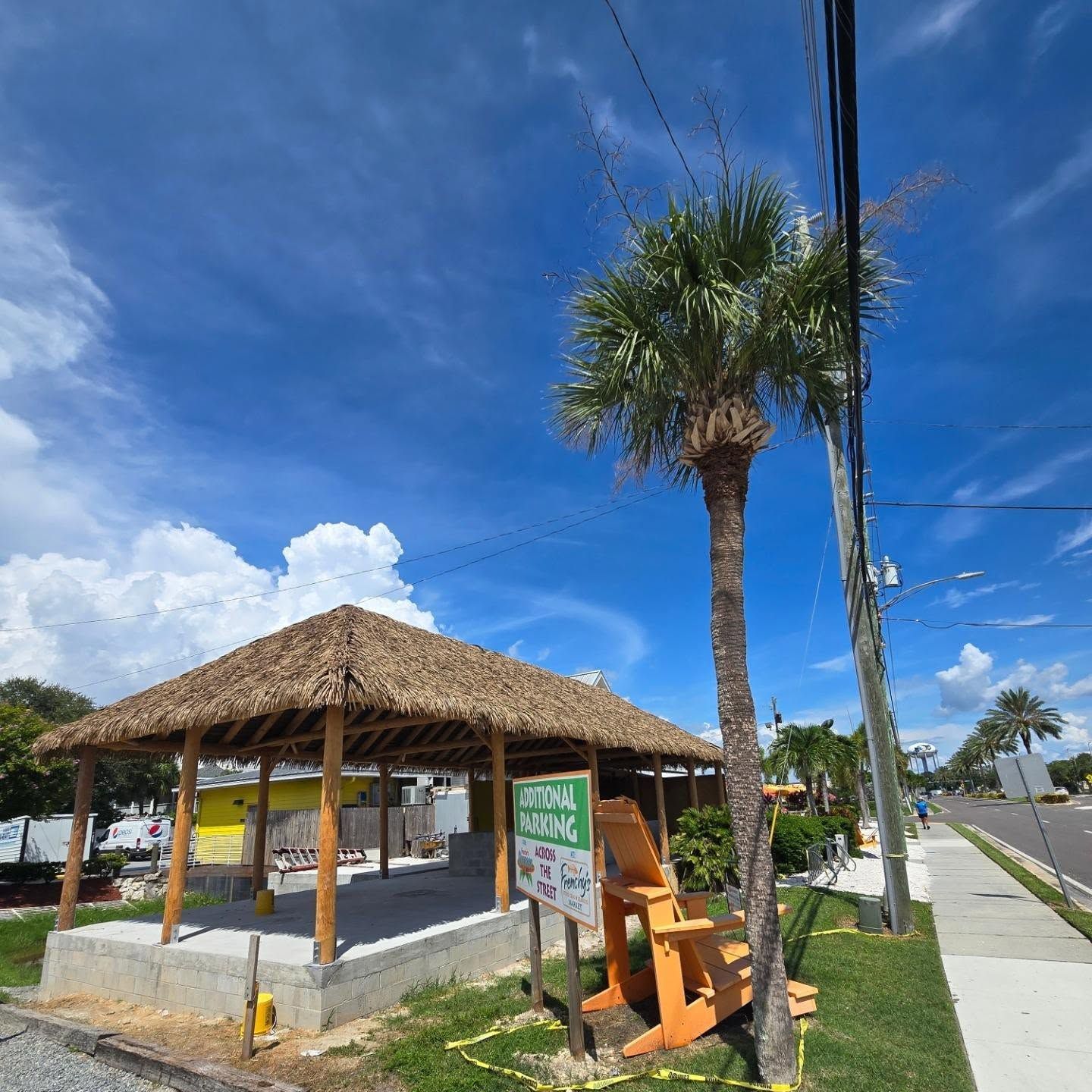 Tiki hut with thatched roof, palm tree, and wooden chair on a sunny street.