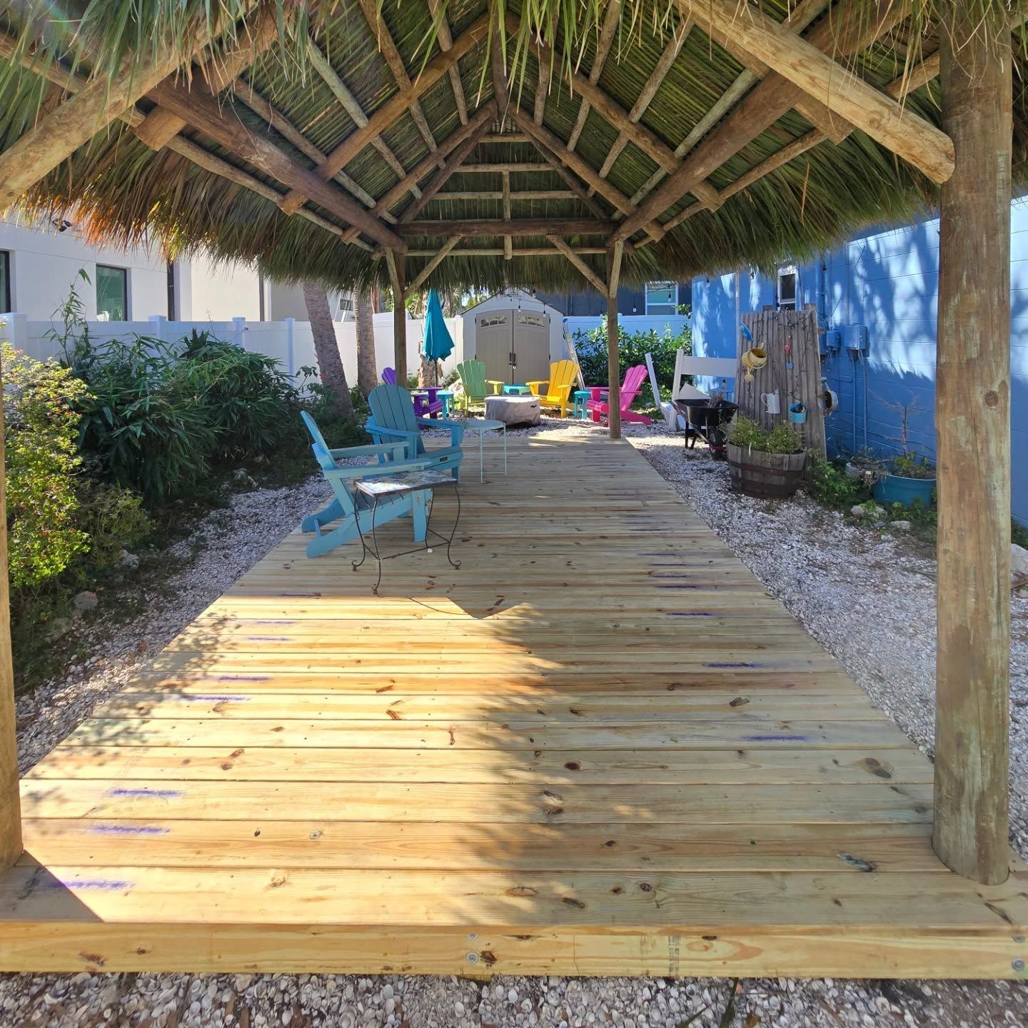 Wooden walkway under thatched roof, leading to colorful chairs and yard.