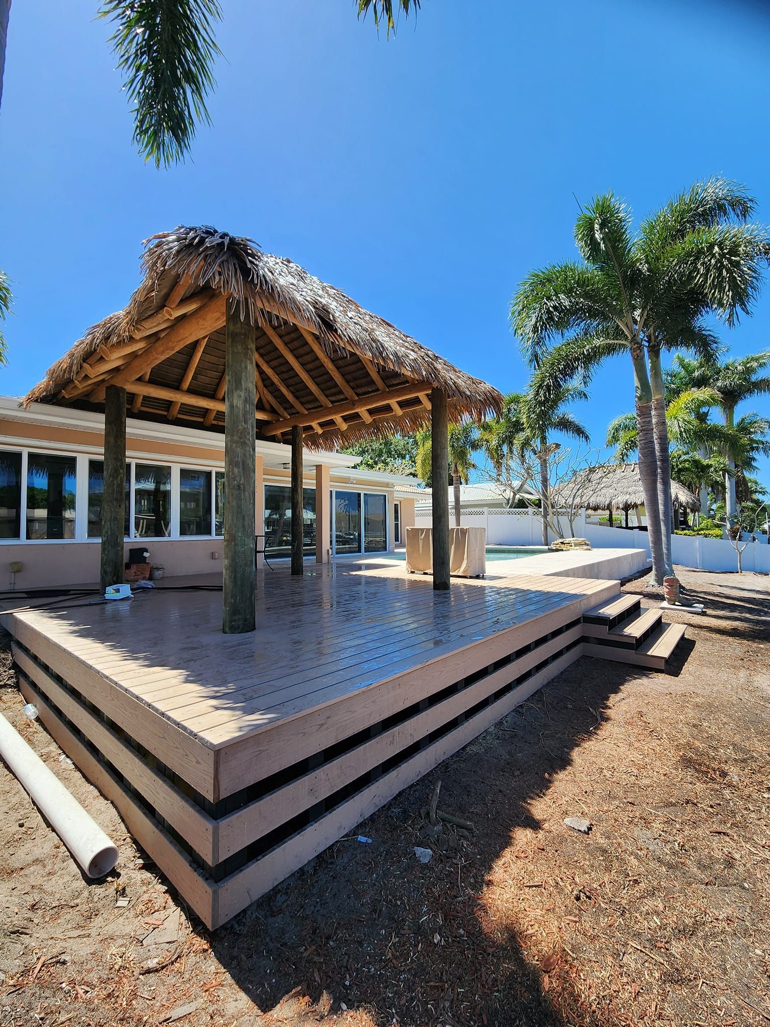 Wooden tiki hut on a raised concrete patio, next to a building with windows, under a blue sky.