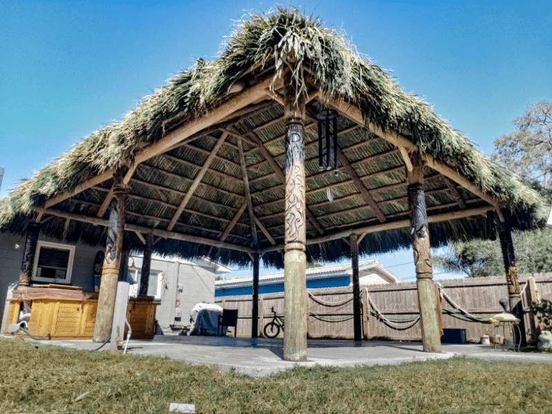 Tiki hut with thatched roof, wooden beams, and poles, set on concrete patio with green grass in sunny outdoor space.