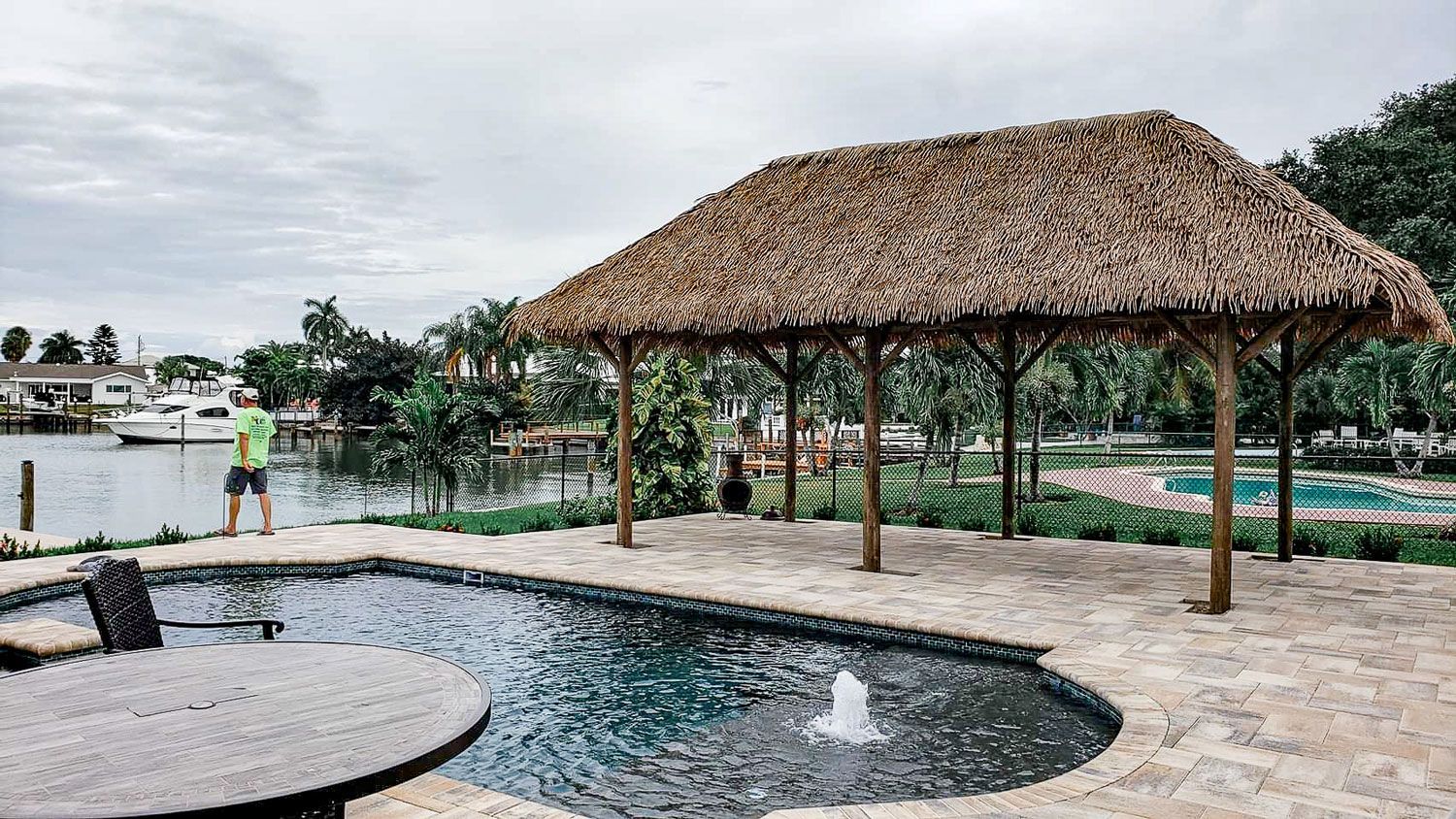 Poolside cabana with thatched roof, man fishing, water, and cloudy sky.