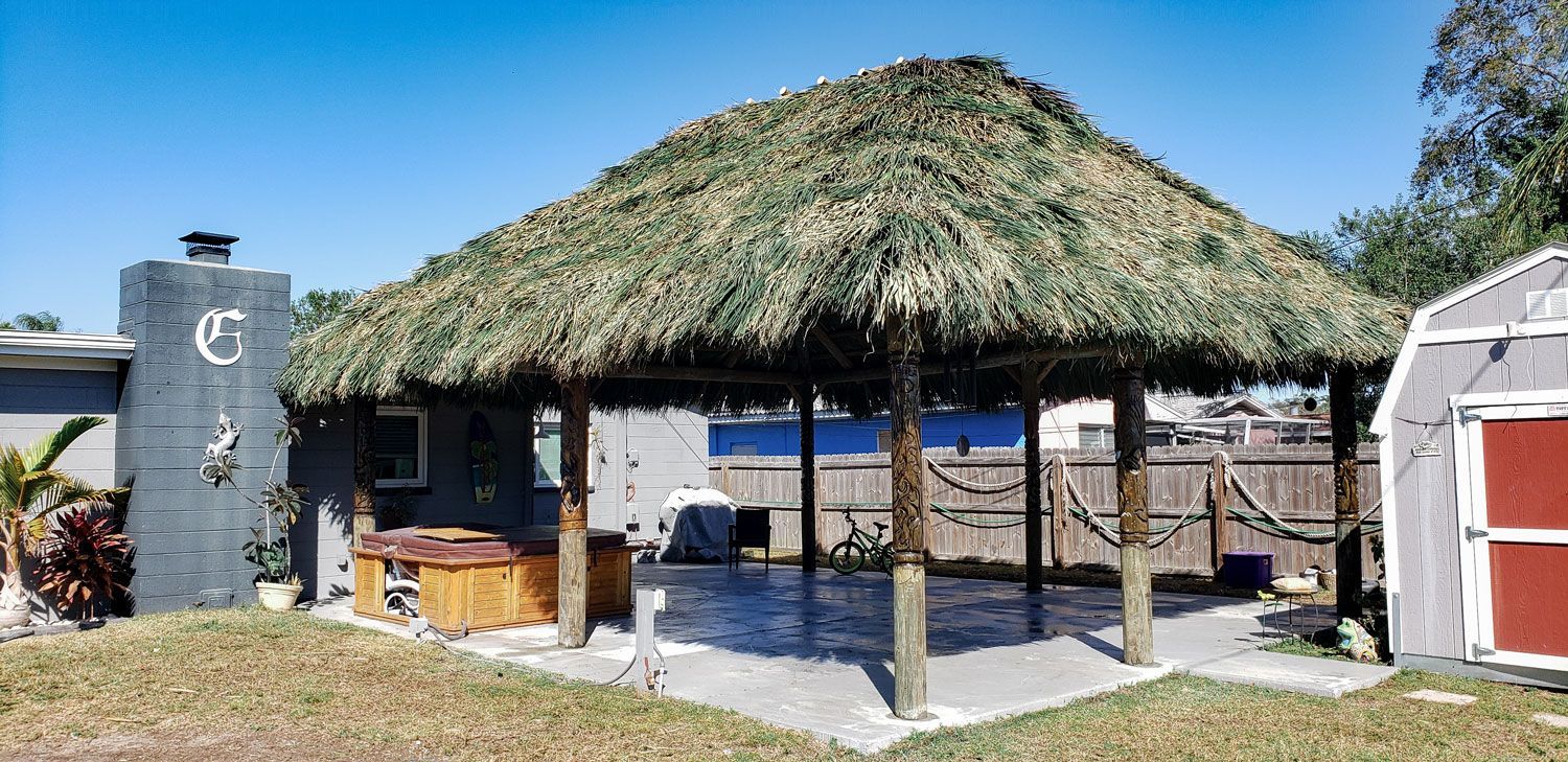 Thatched-roof tiki hut with wooden supports, next to a building and a shed, on a sunny day.