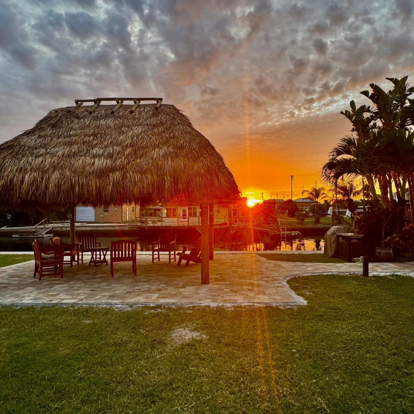 Sunset over a thatched-roof cabana by a waterway with a grassy lawn.