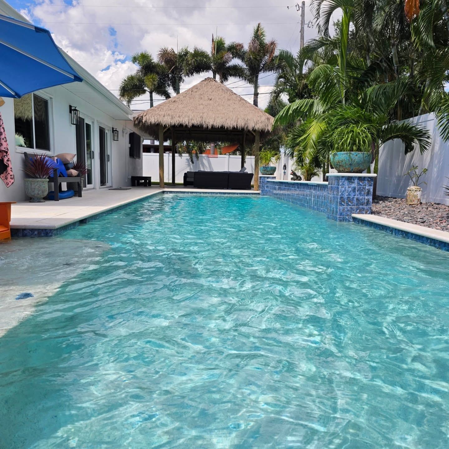 Pool in tropical setting; white house, thatched roof cabana, palm trees, blue water.