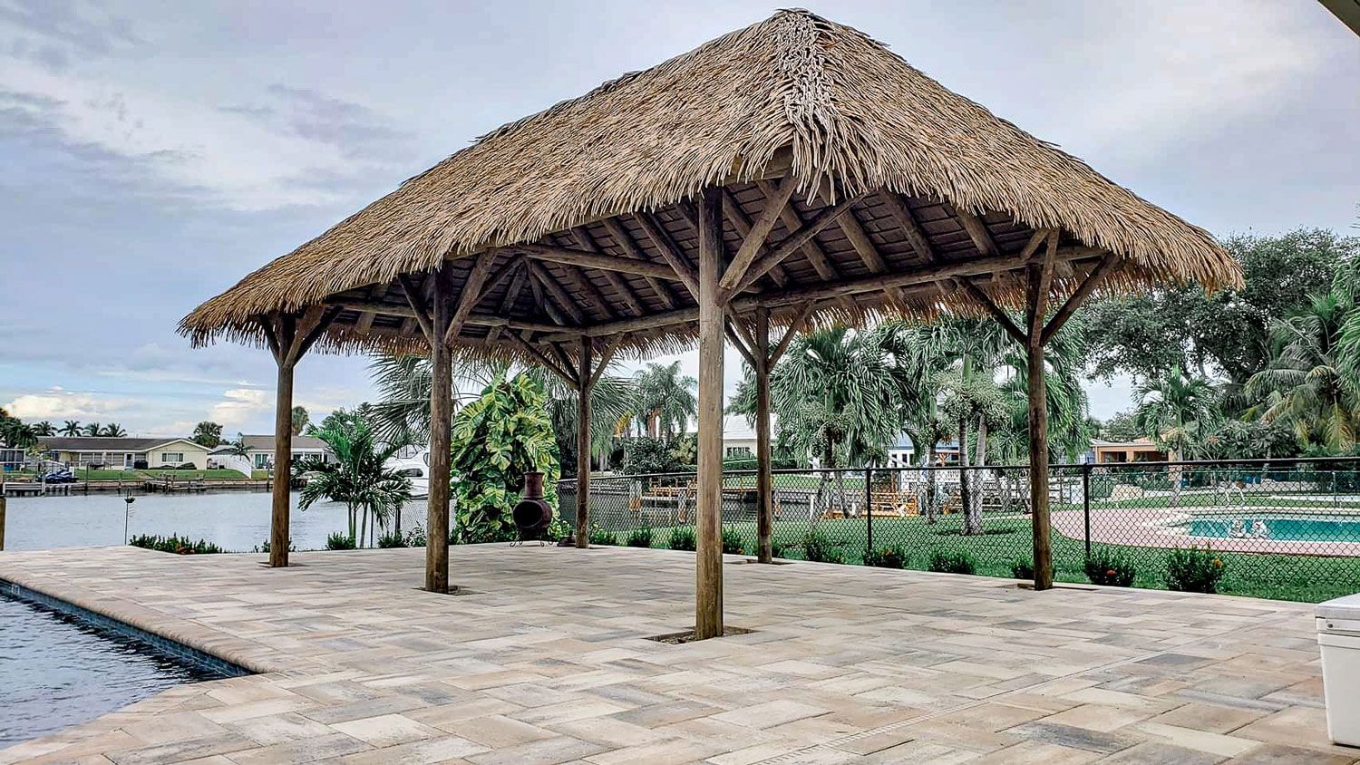 Tiki-style roofed gazebo on a patio, overlooking water and greenery.