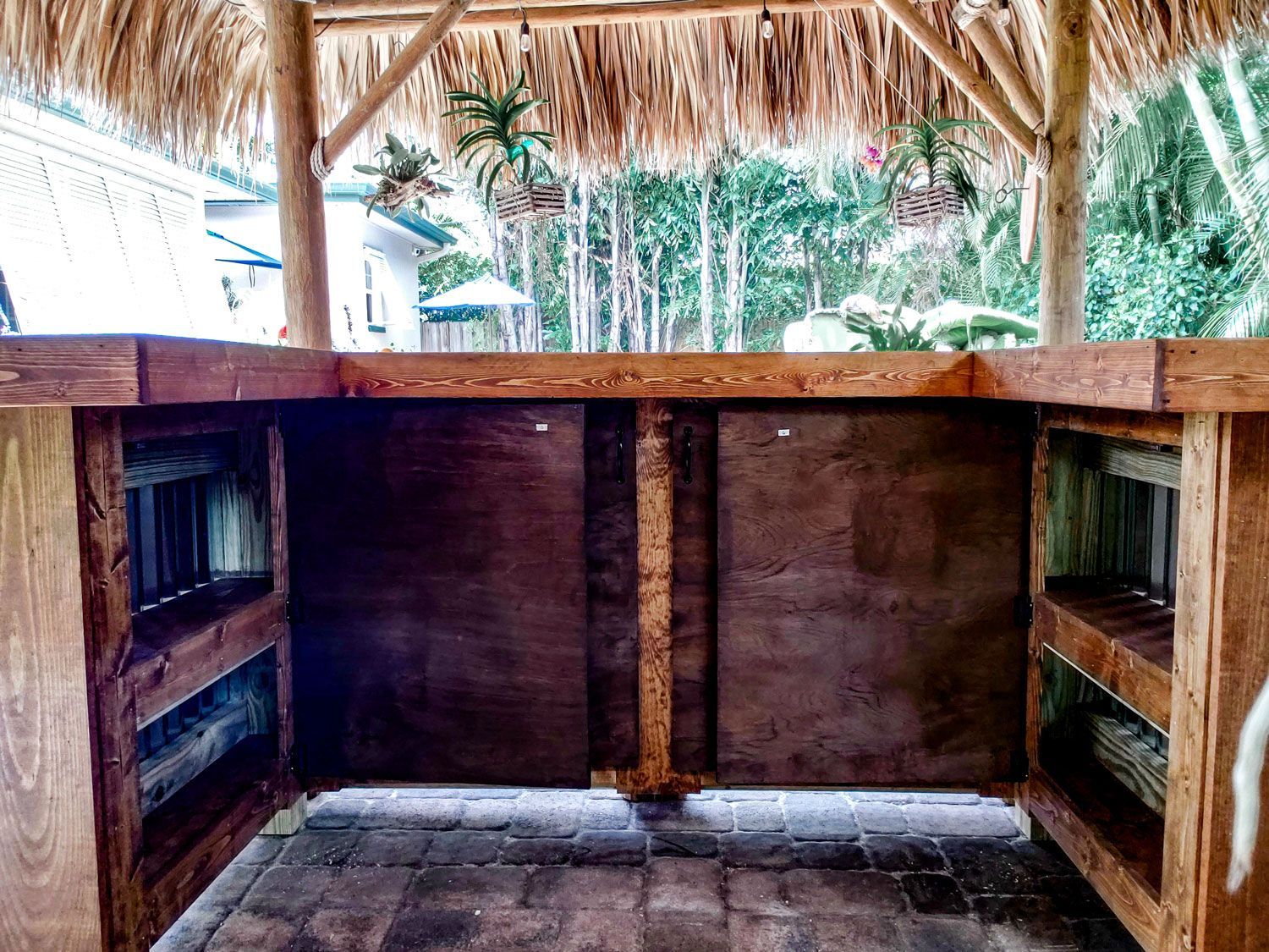 Outdoor tiki bar with thatched roof, wooden structure, shelves, and dark brown backing.
