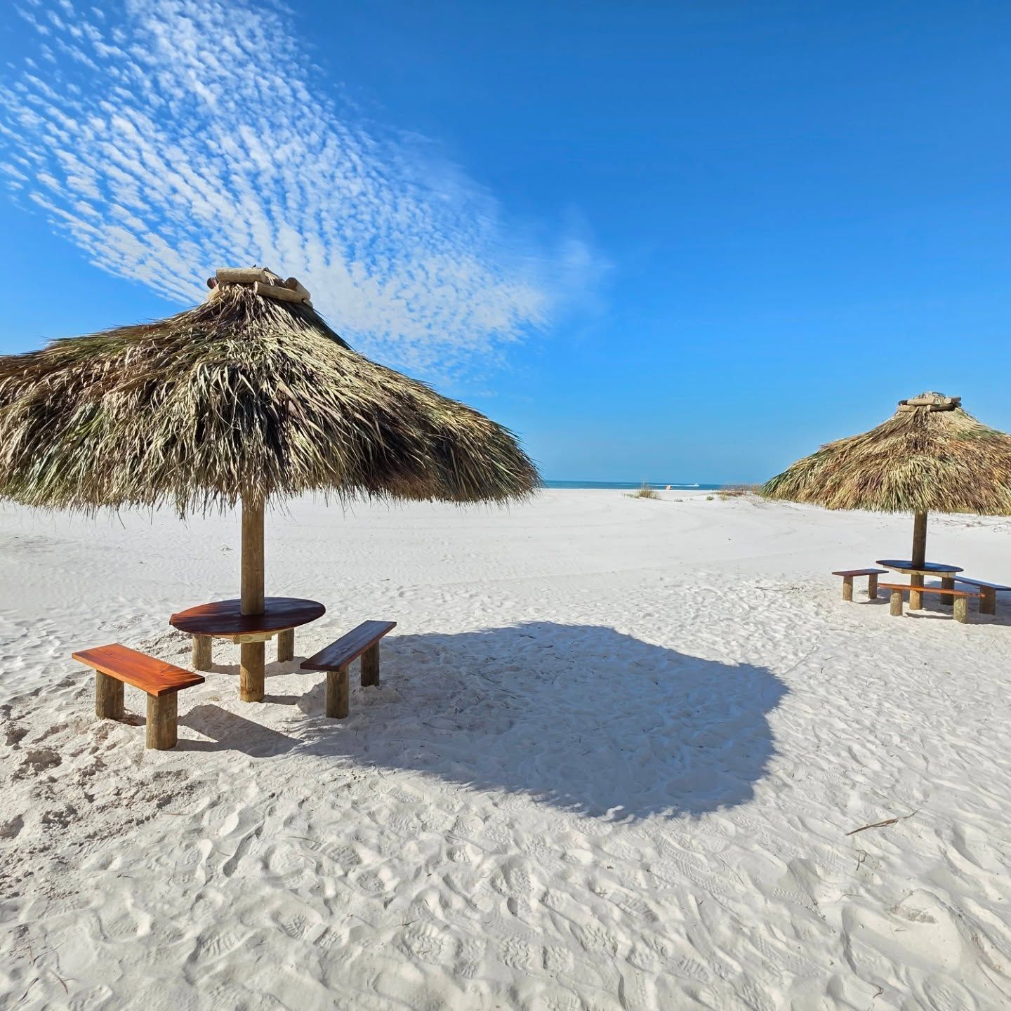 Two thatched umbrellas on a white sand beach with picnic tables, under a blue sky.