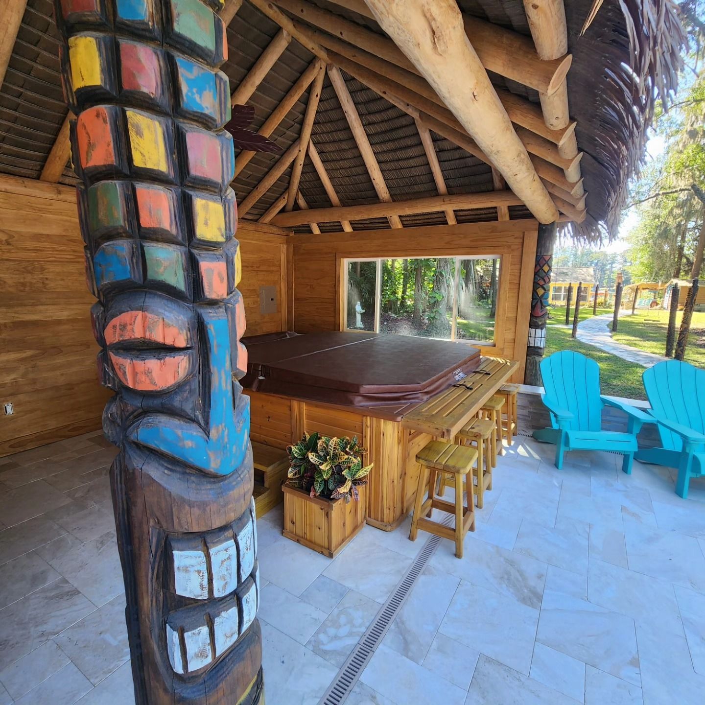 Tiki bar with hot tub under thatched roof; colorful totem pole, turquoise chairs, and stools.