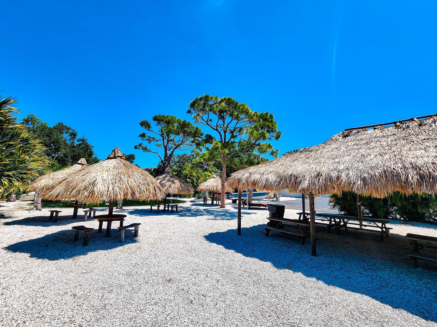 Outdoor dining area with thatched umbrellas, picnic tables, and white gravel. Bright blue sky.