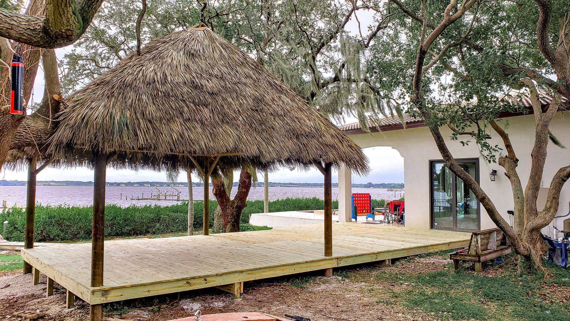 A thatched roof gazebo on a wooden deck overlooking a body of water and foliage.