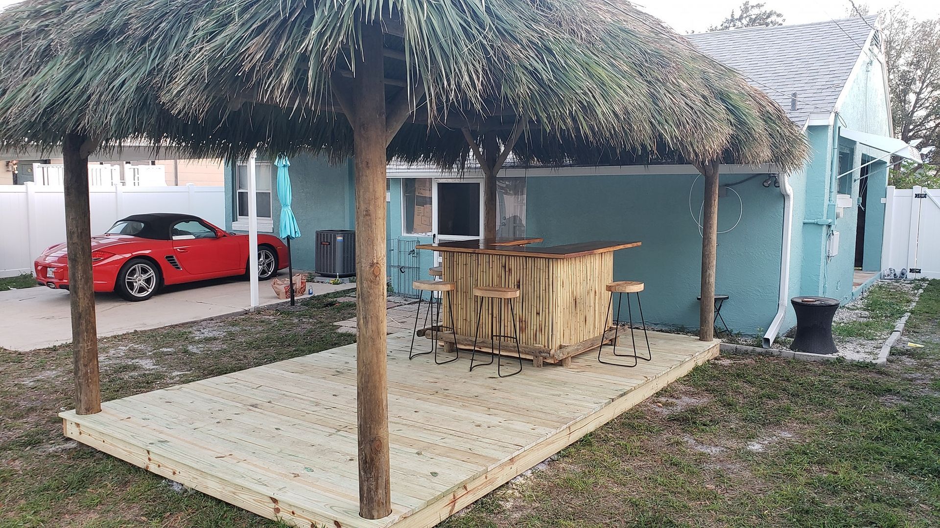 Tiki bar with thatched roof and wooden deck in backyard; red car in background.