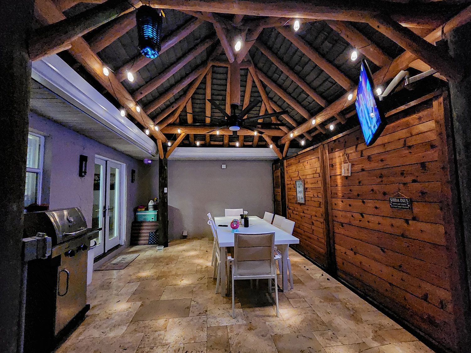 Outdoor dining area under a wooden roof with a grill, table, TV, and string lights.