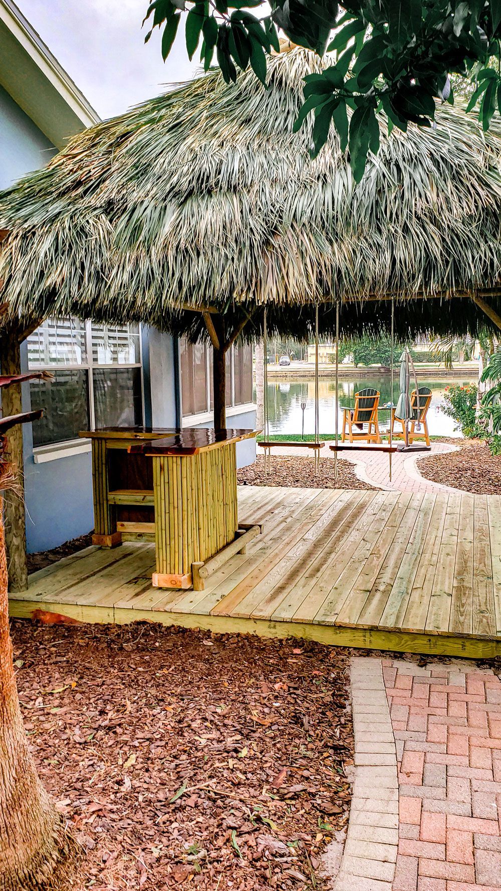 Wooden deck with thatched roof gazebo, overlooking a body of water. Brick pathway in foreground.