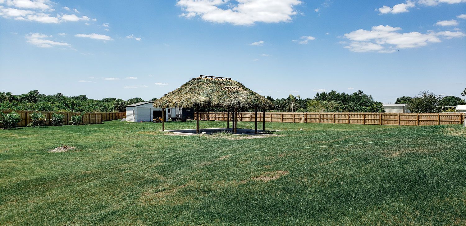 A thatched-roof structure stands in a grassy yard under a blue sky, surrounded by a wooden fence and trees.
