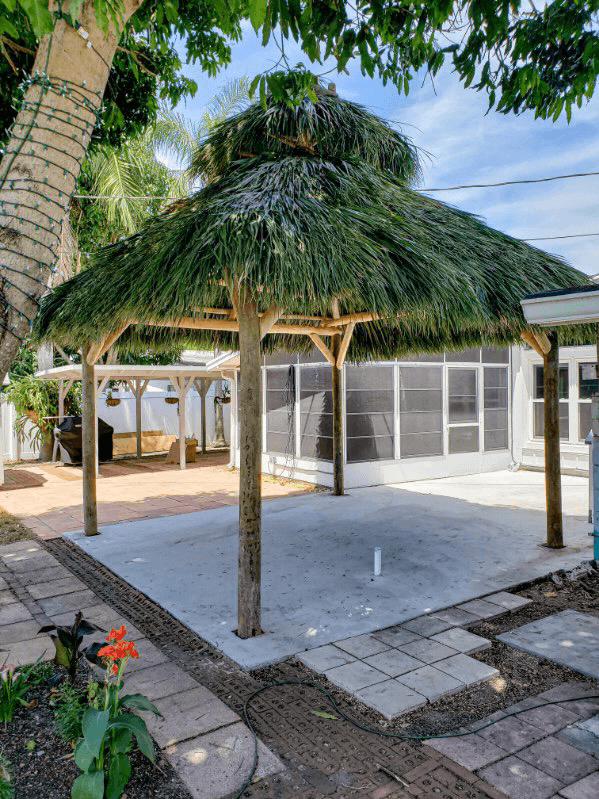 Tiki hut with layered thatched roof on concrete patio, surrounded by brick walkway and screen enclosure.